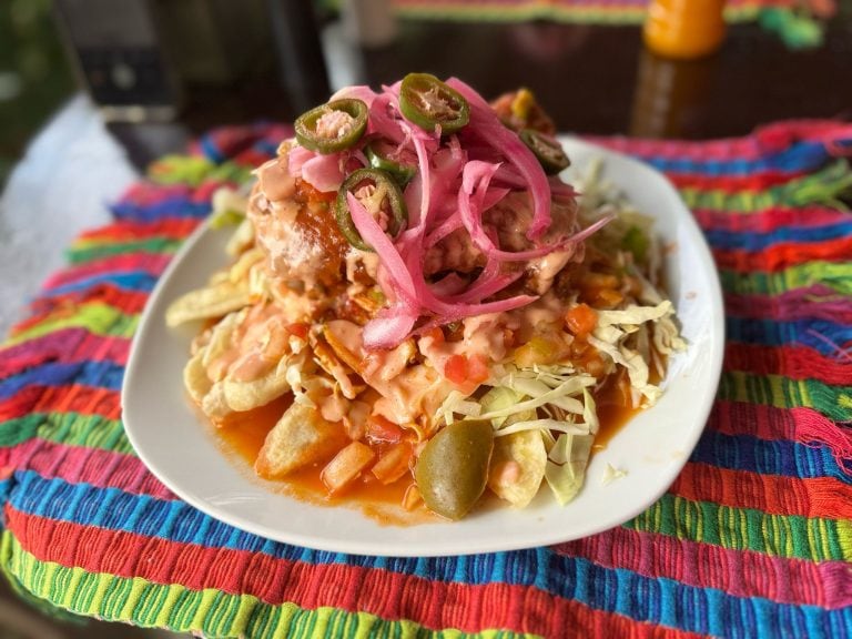 A plate of traditional Honduran tacos topped with jalapeños, shredded cabbage, and pink pickled onions is placed on a vibrant, multicolored woven cloth in Roatan, Honduras.