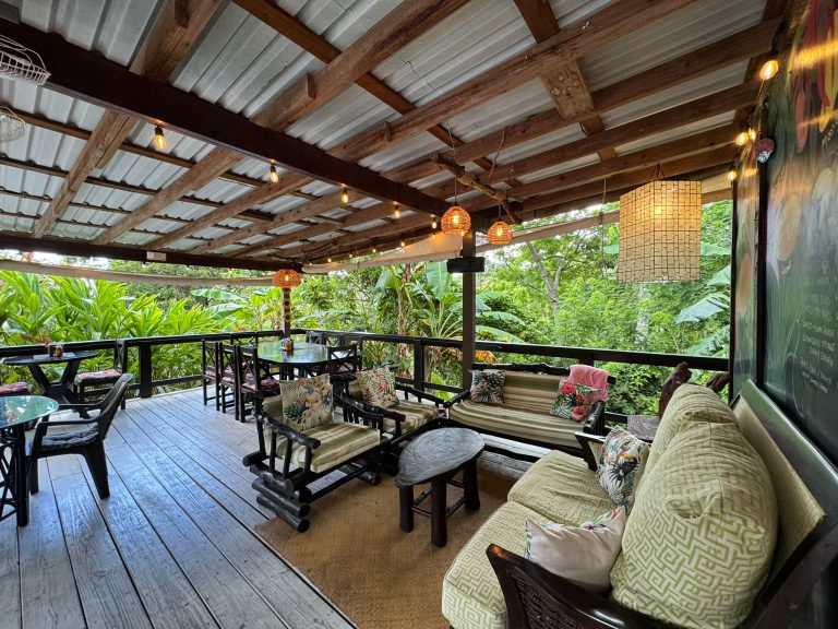 An open-air restaurant or bar in Roatan, Honduras, featuring wooden furniture, patterned cushions, hanging wicker light fixtures, and surrounded by lush green tropical foliage.