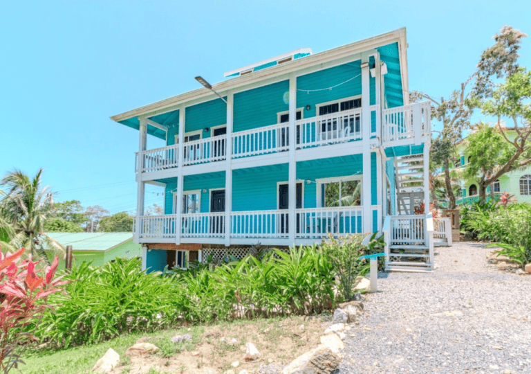 A three-story turquoise house with white railings and stairs is situated among lush tropical plants on Roatan, Honduras, with a gravel pathway leading to the entrance and other colorful buildings visible in the background.