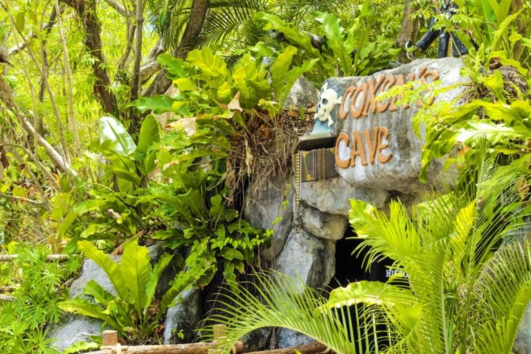 A large rock sign reading "COVENANT CAVE" with a skull decoration and string beads, surrounded by lush green tropical plants and trees in a dense, vibrant forest setting in Roatan, Honduras.