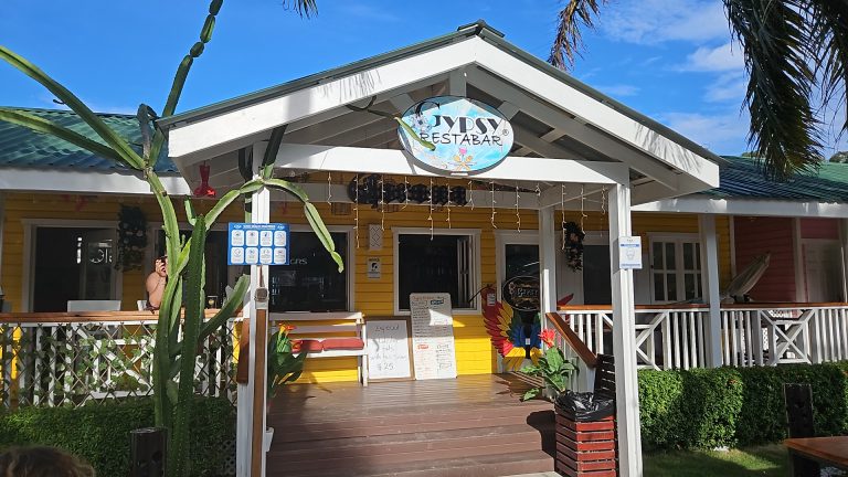 A colorful yellow building with a white peaked roof and a sign reading "Gypsy Restabar" is situated on Roatan, Honduras, featuring a wooden deck, tropical plants, a cactus, and a person taking a photo in front of the entrance under a clear blue sky.