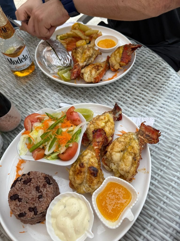 A person with a watch is serving a seafood dish with grilled lobster tails, fried fish, and side salads on a white plate at a restaurant in Roatan, Honduras, with small bowls of dipping sauces, lime wedges, and a bottle of Modelo beer visible on a wicker table.