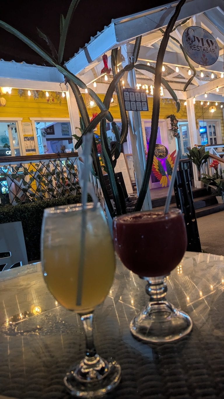 Two colorful drinks in cocktail glasses are placed on a table outside a brightly lit restaurant in Roatan, Honduras, with the restaurant's yellow exterior, string lights, and tropical decor visible in the background.