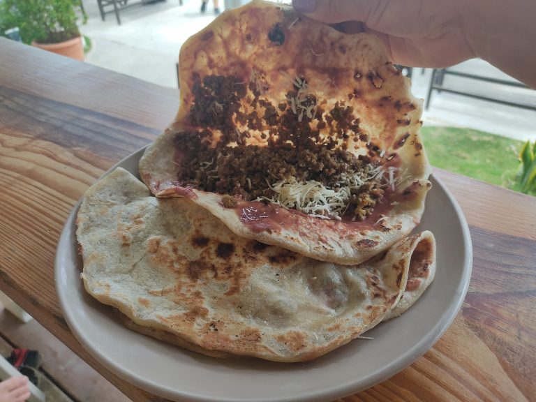 A plate of tacos with seasoned meat, cheese, and toppings sits on a wooden table near a balcony with a view of greenery in Roatan, Honduras.