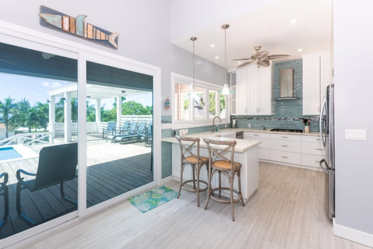 Modern kitchen inside a house in Roatan Honduras with white cabinets, a blue backsplash, and a sliding glass door leading to an outdoor deck with lounge chairs and tropical greenery.
