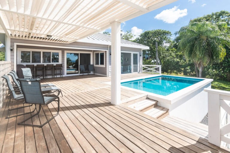 A wooden outdoor deck with a small swimming pool, surrounded by tropical trees and a white house on Roatan, Honduras, featuring sliding glass doors and windows.