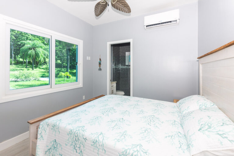 A cozy bedroom in Roatan, Honduras, featuring a large window overlooking lush tropical trees and greenery outside, with a white bed covered in seabed coral patterned bedding, and an air conditioning unit mounted on the light gray wall.