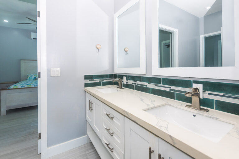 Modern white double vanity bathroom with two large mirrors, green and white tile backsplash, and a view into a bedroom with a bed and tropical bedding on Roatan Honduras.