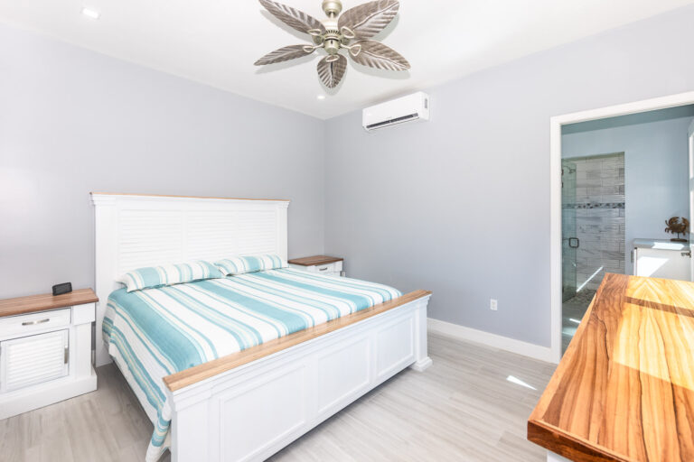 A bedroom in Roatan, Honduras, featuring a white wooden bed frame with a blue and white striped bedspread, two nightstands with wooden tops, and a wooden dresser against a light gray wall.