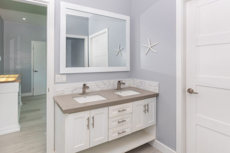 Modern double vanity bathroom with two sinks, a large white-framed mirror, and decorative starfish on the light grey wall in a coastal-style home in Roatan, Honduras.