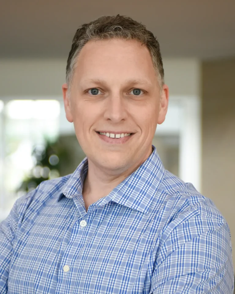 Close-up of a smiling man with short, light brown hair and blue eyes wearing a blue checkered shirt in an indoor setting in Roatan, Honduras.