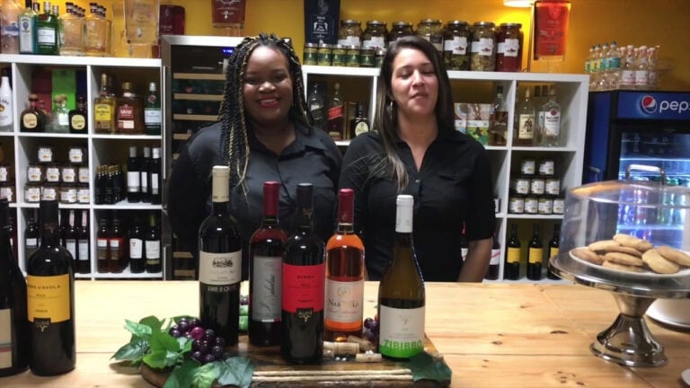 Two women stand behind a wooden counter in a wine shop in Roatan, Honduras, with shelves stocked with bottles of wine, liquor, and jars of preserves in the background.