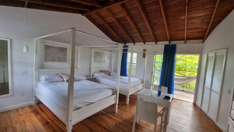 A cozy bedroom in Roatan, Honduras features two white four-poster beds with white linens, wooden floors, large windows with blue curtains, and a balcony with a hammock overlooking lush green foliage.