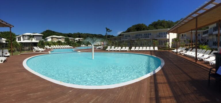 A resort pool area in Roatan, Honduras features a large, freeform swimming pool surrounded by white lounge chairs, palm trees, and multi-story white buildings with balconies set against lush green hills and a clear blue sky.