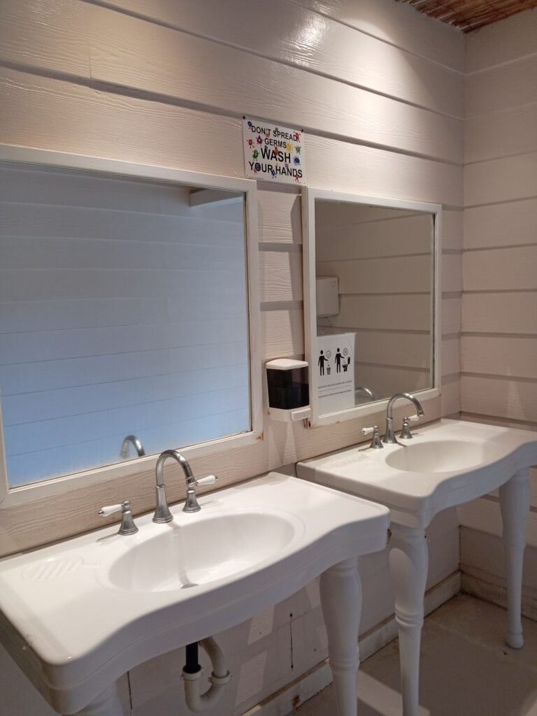 A row of three white sinks with faucets and mirrors above, located in a restroom with wood-paneled walls in Roatan, Honduras, featuring a sanitizer dispenser and a sign reminding to wash hands to prevent the spread of germs.