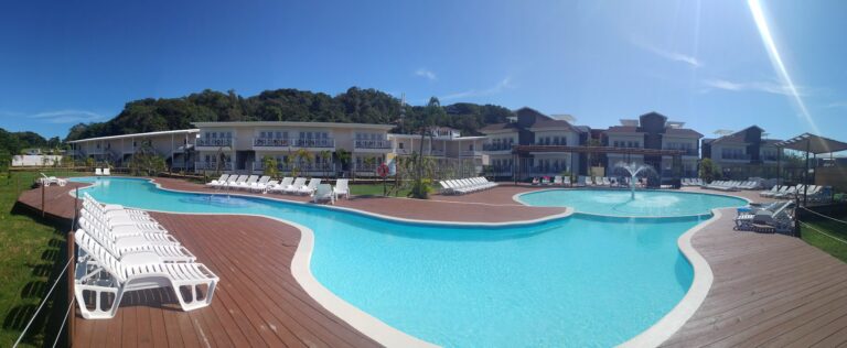 A large resort pool area surrounded by white sun loungers and multi-story buildings with balconies, set against a backdrop of green hills on Roatan, Honduras.