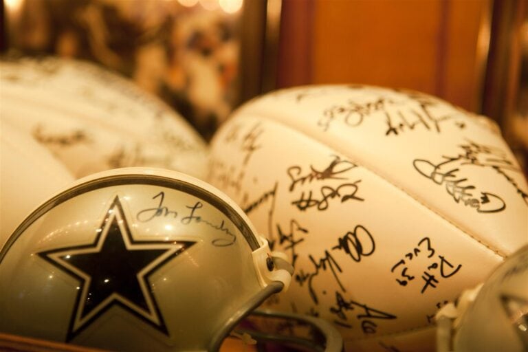 A white Dallas Cowboys football helmet signed by various players is displayed on a surface, with illuminated signed footballs in the background, in a setting related to Roatan, Honduras.