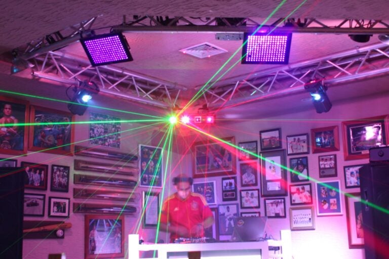 A DJ performs with colorful laser lights on the ceiling of a nightlife venue in Roatan, Honduras, surrounded by framed photos and artwork on the walls.