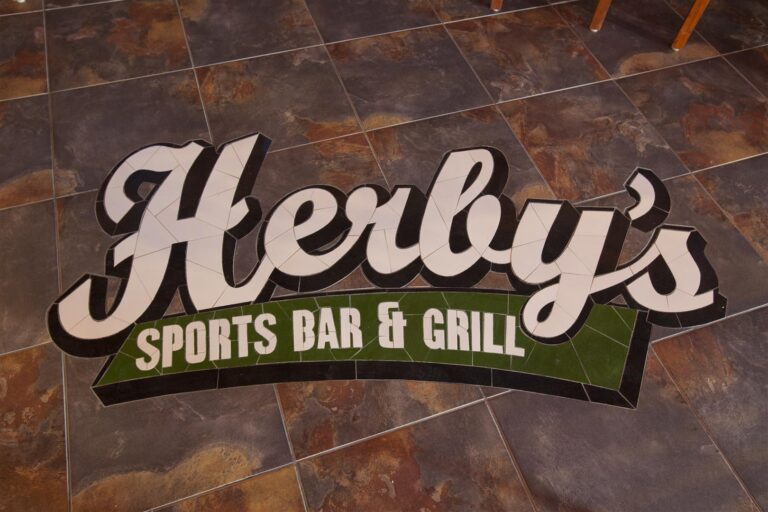 A colorful sign on tiled flooring for "Herley's Sports Bar & Grill" in Roatan, Honduras, featuring large cursive lettering and a green banner with the establishment's name and type.
