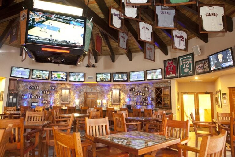 A sports bar in Roatan, Honduras, featuring multiple flat-screen TVs displaying various games, wooden tables and chairs, a stone-walled bar area with liquor bottles, and hanging sports jerseys from different teams in the ceiling.