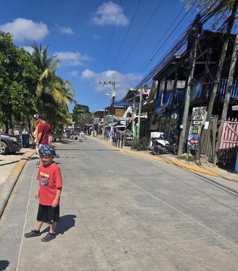 A boy wearing a red shirt and black shorts walks on a sunny street in Roatan, Honduras, lined with shops, palm trees, and utility poles under a bright blue sky with scattered clouds.
