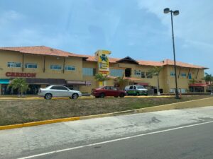 A Hospital Hernandez in Roatan, Honduras, features a yellow building with a colorful butterfly and flower logo on its signage, situated along a busy street with parked cars and a clear blue sky overhead.