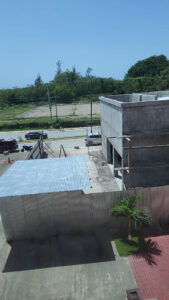 A view of a construction site next to a busy street in Roatan, Honduras, with an unfinished concrete building, a corrugated metal roof structure, parked cars, and a small palm tree, against a backdrop of green trees and a clear blue sky.