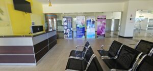 An indoor waiting area at a medical facility in Roatan, Honduras, featuring black chairs, a reception desk, informational banners, and glass doors leading to medical offices.