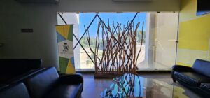 An interior view of a waiting area at Roatan Hospital in Honduras, featuring black leather sofas, a glass-top table with a decorative wooden sculpture, a colorful Hospital Cemesa Roatan banner, and large windows revealing a clear blue sky and greenery outside.