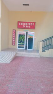 A hospital entrance with signs indicating 24-hour emergency services and an elevator, located in Roatan, Honduras.
