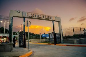 The entrance to the Satuye Hospital in Roatan, Honduras, features a large white sign with the hospital's name, a security gate, a blue parking area sign, and a sunset sky in the background.
