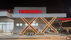 A brightly lit emergency room entrance in Roatan, Honduras, features a large red "EMERGENCIA" sign above the doorway, with a hospital parking area and motorcycles parked outside at night.
