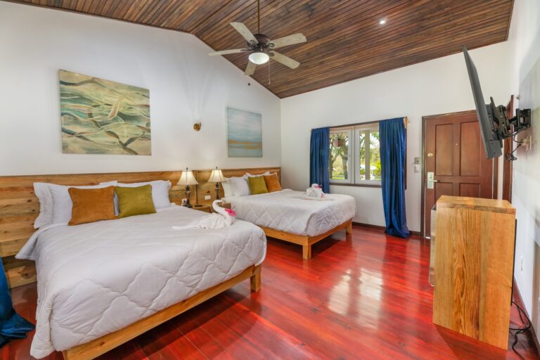 A bedroom in a hotel or vacation rental on Roatan, Honduras, features two neatly made beds with white linens, colorful pillows, wooden headboards, a ceiling fan, artwork on the white walls, a window with blue curtains, and hardwood floors.