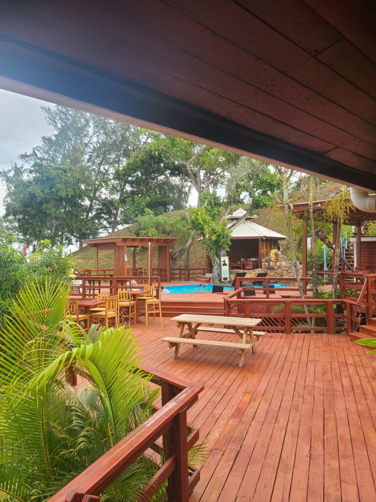 A wooden deck with picnic tables and seating areas overlooks a lush tropical garden with trees and plants in Roatan, Honduras, featuring a pool and gazebo in the background.