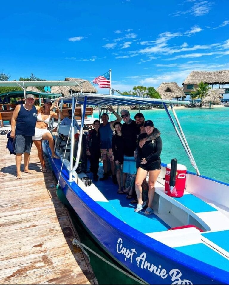 A group of tourists stands on a wooden dock next to a blue and white boat named "Captain Annie Boy" at a tropical waterfront in Roatan, Honduras, with thatched-roof huts, lush greenery, and the American flag flying against a bright blue sky in the background.
