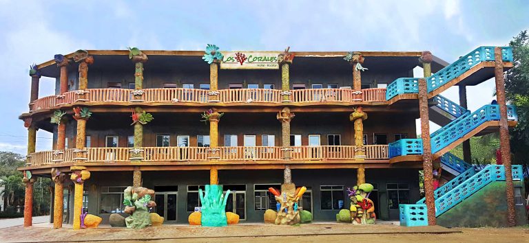 The three-story hotel "Los Corales" in Roatan, Honduras, features vibrant, colorful decor with coral and sea-themed sculptures, a large sign at the top, and bright blue outdoor staircases on the right side.