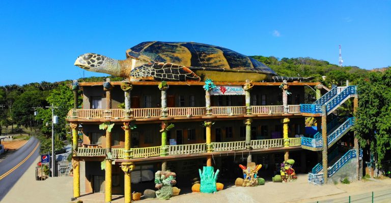 A colorful, multi-story hotel in Roatan, Honduras, features a giant sea turtle sculpture on the roof and vibrant tropical decor with marine-themed ornaments and bright blue stairs.