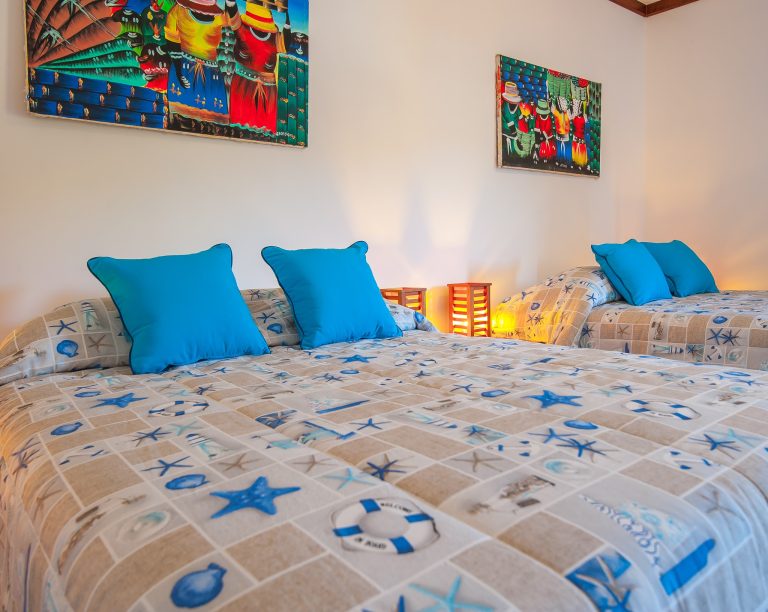 A bedroom in a Roatan, Honduras accommodation features a bed with a starfish-themed bedspread and multiple blue pillows, two lit lanterns on the bedside table, and colorful artwork hanging on the white wall above.