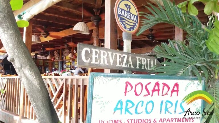 A view of a tropical-style bar and restaurant in Roatan, Honduras, featuring wooden decor, ceiling fans, colorful signage including "Cervezas Frias" and "Posada," with lush green plants and a glimpse of the interior's bar area with bottles and glasses.