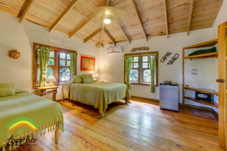A cozy bedroom in Roatan, Honduras, features wooden floors, a vaulted wooden ceiling, two beds with green bedspreads, green curtains on windows, and colorful decorative art on the walls.