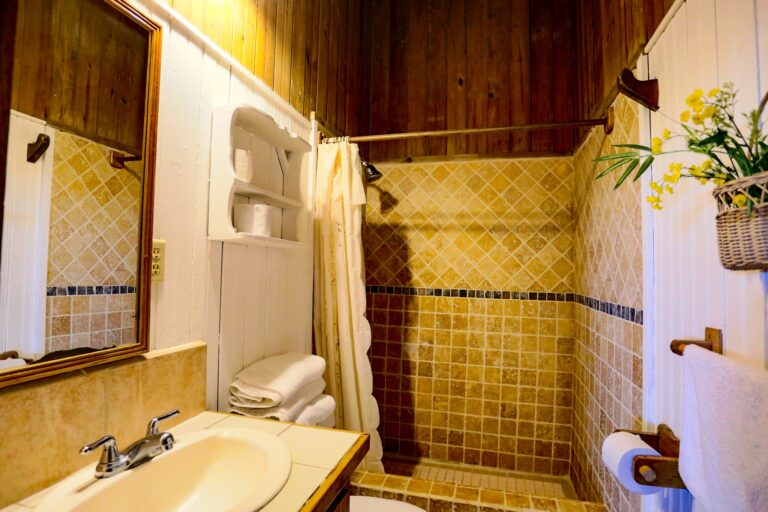 A cozy bathroom in a Roatan, Honduras accommodation features a wooden ceiling, tiled walls with a diamond pattern, a white vessel sink with a chrome faucet, a mirror framed in wood, and neatly folded white towels with a colorful flower arrangement on the side.