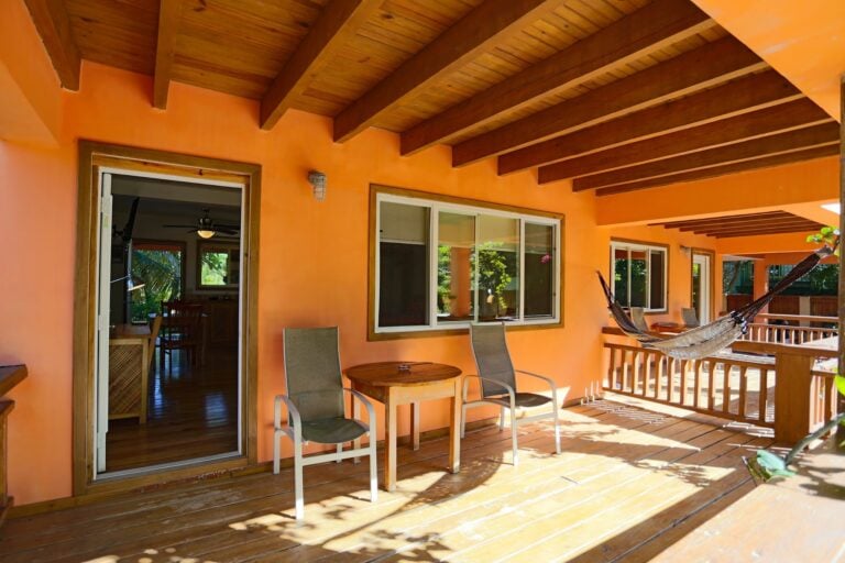 A covered outdoor patio with orange walls, wooden ceiling beams, sliding glass doors, chairs, and a hammock, located in Roatan, Honduras.