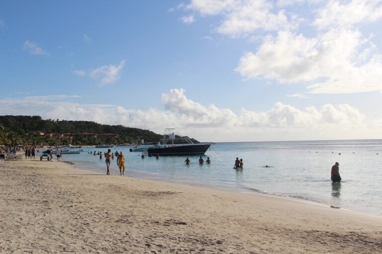 A sandy beach in Roatan, Honduras, features people swimming and relaxing near the clear ocean waters with a large boat anchored offshore and a lush green hillside in the background under a partly cloudy sky.