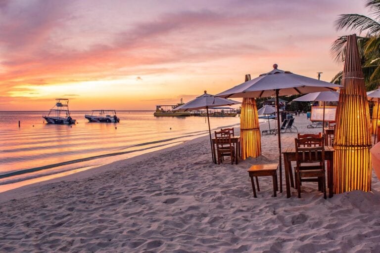 Colorful sunset over Roatan's sandy beach with boats anchored in calm waters, surrounded by white umbrellas, wooden tables, and palm trees in Honduras.