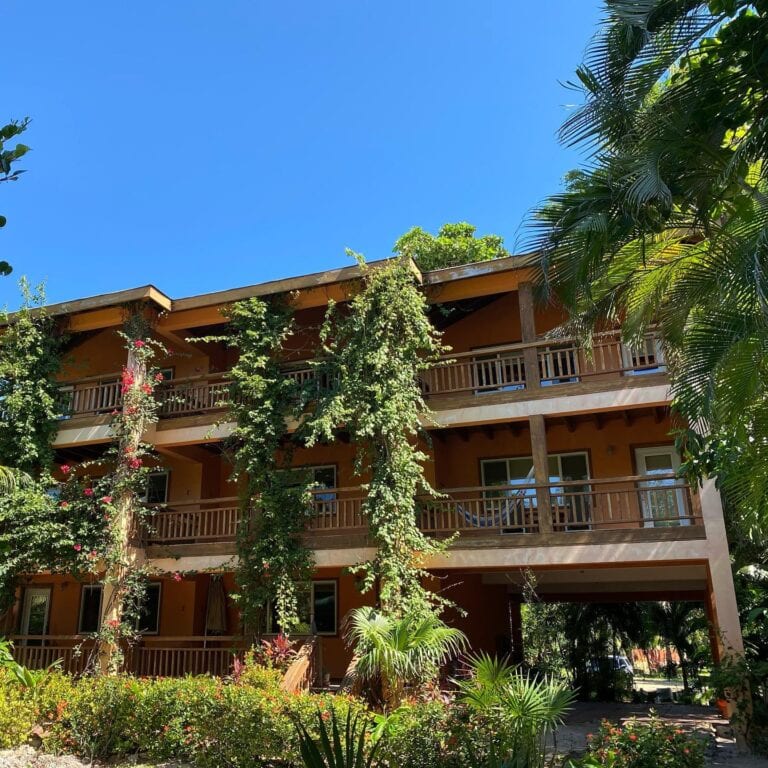 A three-story wooden building with balconies surrounded by lush tropical plants and flowers in Roatan, Honduras, under a clear blue sky.