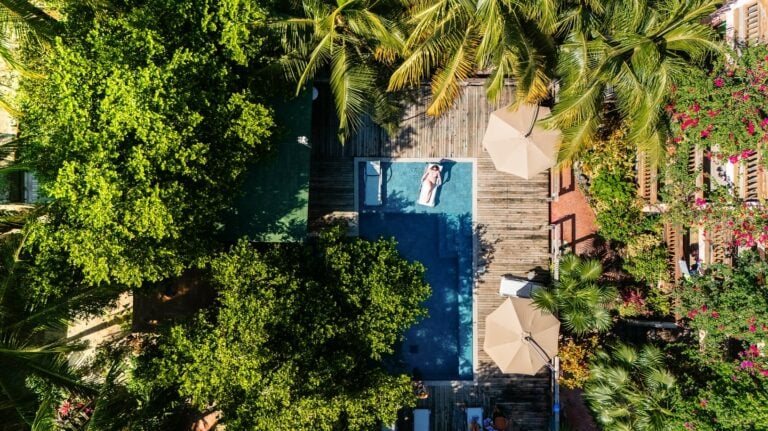 An aerial view of a tropical pool area in Roatan, Honduras, featuring a rectangular swimming pool with a person relaxing on a lounger, surrounded by lush green trees, palm trees, colorful flowering plants, and shaded seating areas with umbrellas.