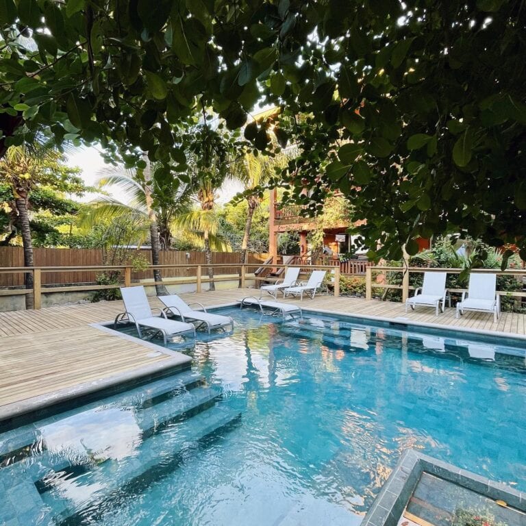 A backyard swimming pool area in Roatan, Honduras, features white lounge chairs on a wooden deck, surrounded by tropical trees and greenery, with a wooden fence and a two-story house visible in the background.