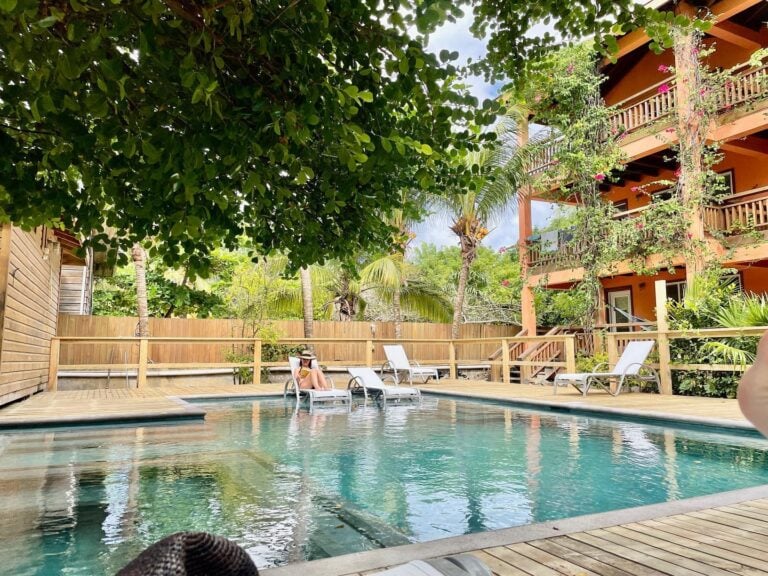 A relaxing pool area with lounge chairs, surrounded by lush tropical trees and a multi-story wooden building with balconies and flowering plants in Roatan, Honduras.