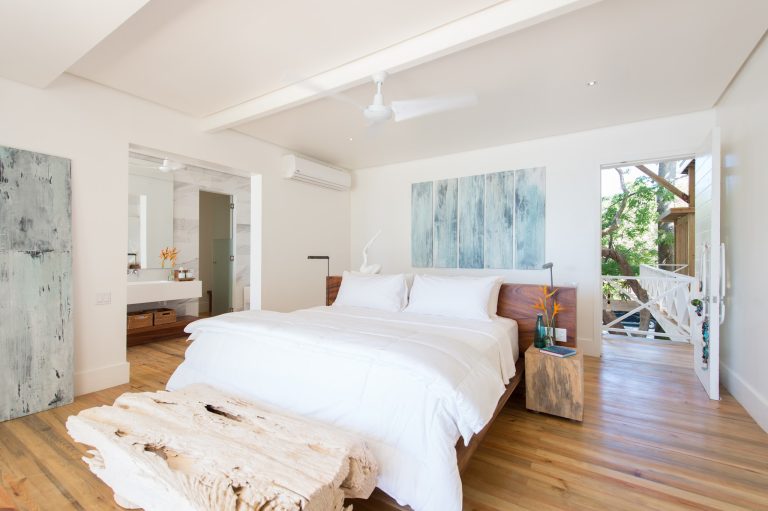 A bright, minimalist bedroom in Roatan, Honduras, features a wooden bed with white linens, a cowhide rug, modern artwork on the walls, and large windows overlooking lush green trees and a small balcony area.