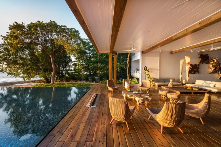 A spacious outdoor patio with wooden decking, modern seating, and decorative wall art overlooks a lush tree and a swimming pool, with a view of the ocean in Roatan, Honduras.